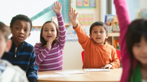 Children sitting in a classroom with their hands raised in the air