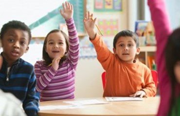 Children sitting in a classroom with their hands raised in the air