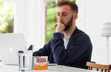 Man with pensive expression sitting at table looking at MacBook with glass of water and box of Mucinex tablets