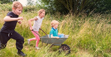 Two children running through a grassy field pushing younger child in wheelbarrow