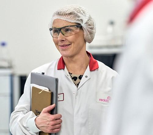 A Reckitt employee in safety glasses, a lab coat and hairnet carries a clipboard