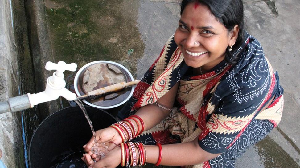 A woman in a sari smiles as she crouches to wash her hands at an outdoor tap