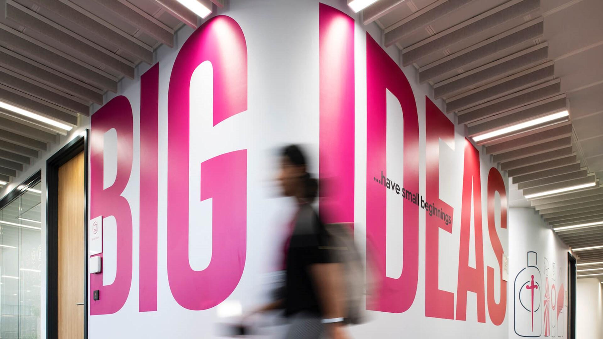 A person is walking through the Reckitt office hallway, past words on the wall that say "Big ideas have small beginnings"