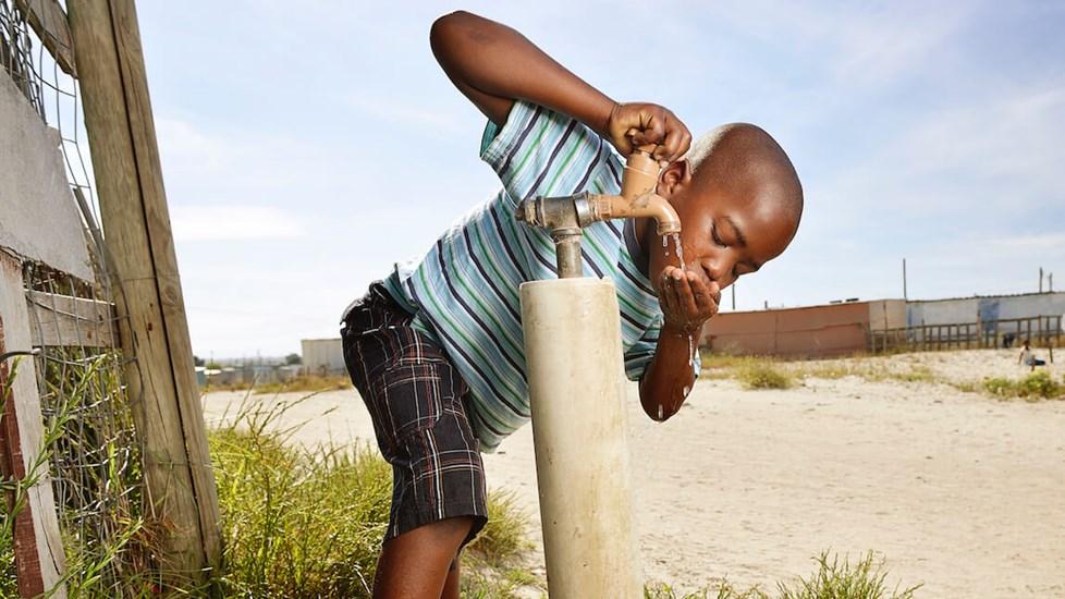 A young African boy drinks water from a tap out of his hands in a barren landscape.