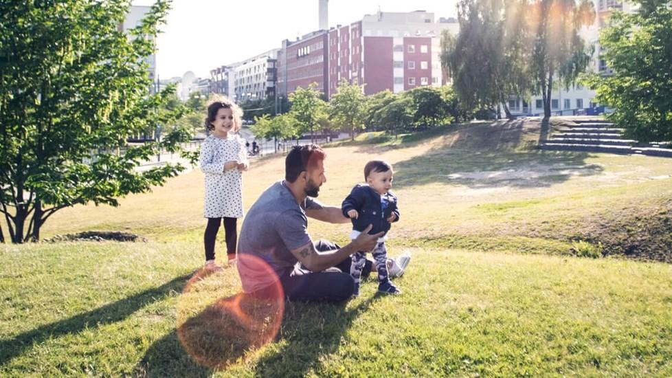 Father playing with children on a grassy field at park against the sky. A cityscape can be seen in the background.